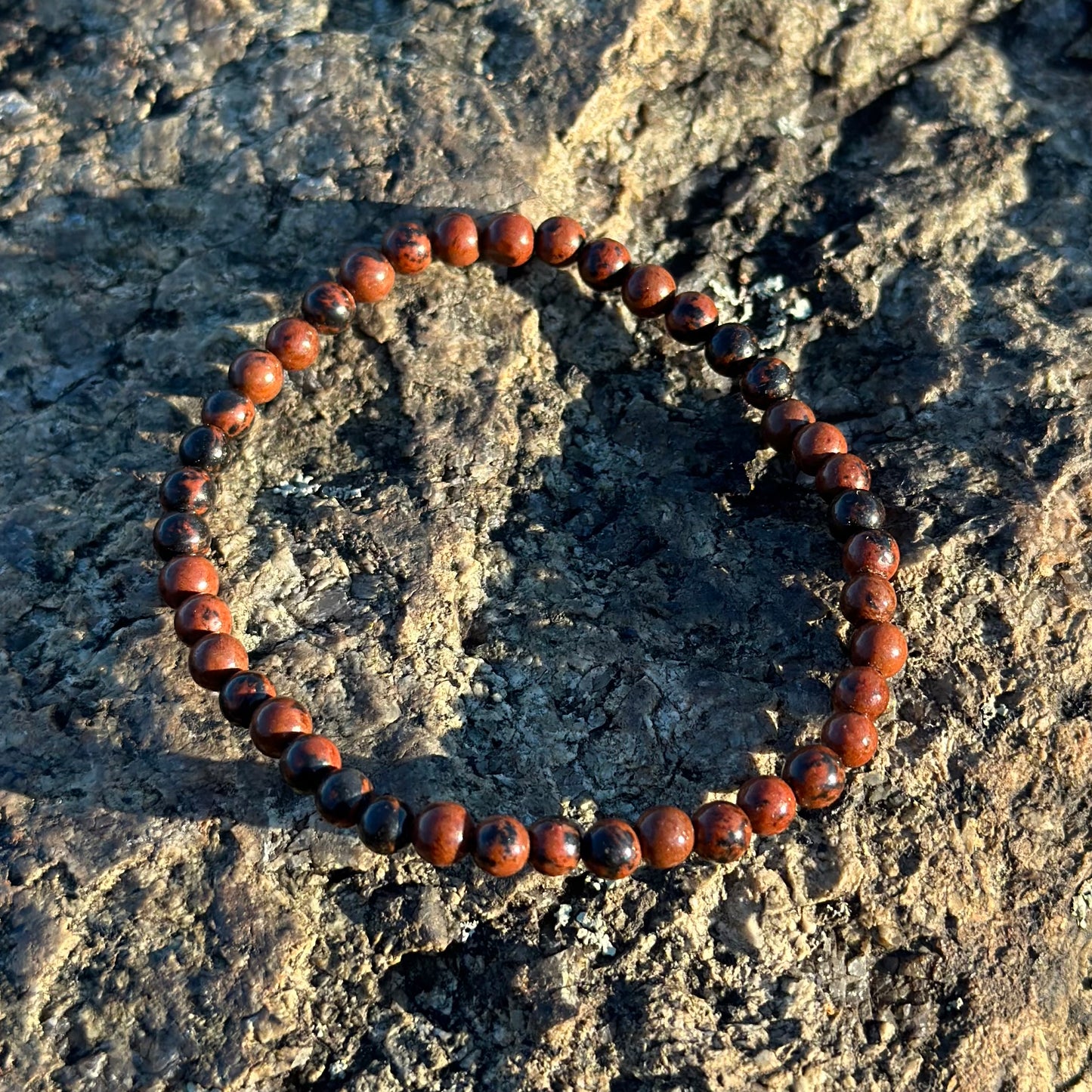 Mahogany Obsidian Bracelet 4MM resting on a textured rock surface, showcasing its polished finish and natural grounding energy.