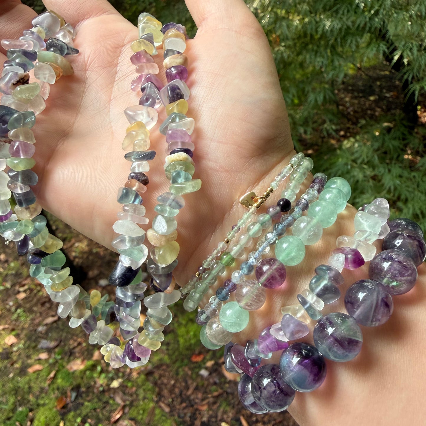A hand holding multiple strands of polished tumbled gemstone beads and several matching beaded bracelets outdoors, with green foliage in the background.