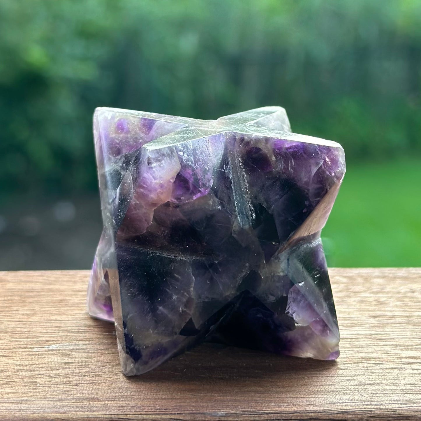 Chevron Amethyst Merkaba crystal sculpture with a blend of purple hues, placed on a wooden surface in a natural outdoor setting.