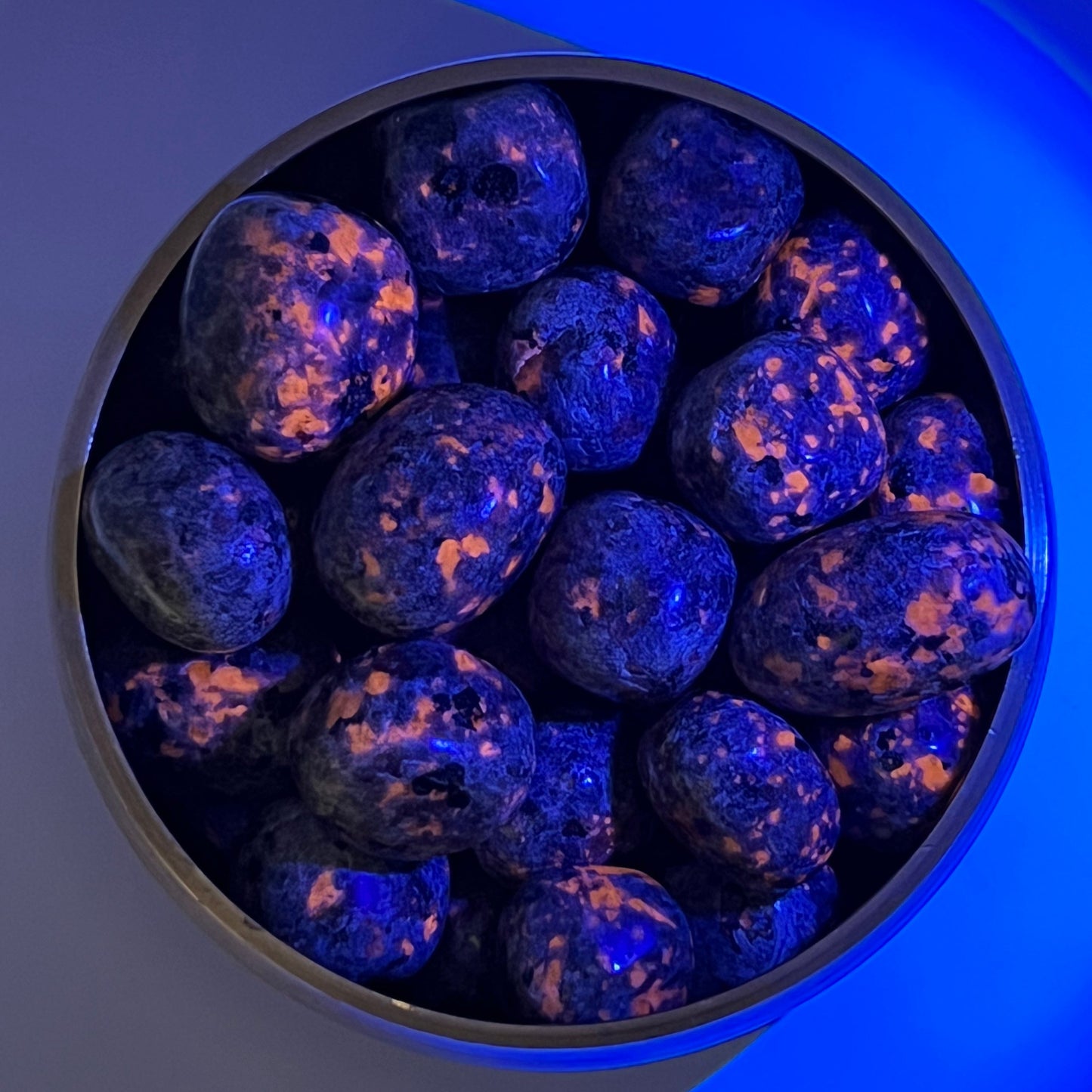 Bowl of fluorescent Larvakite tumbled stones under blue light, showcasing healing properties and sleek dark-grey appearance.